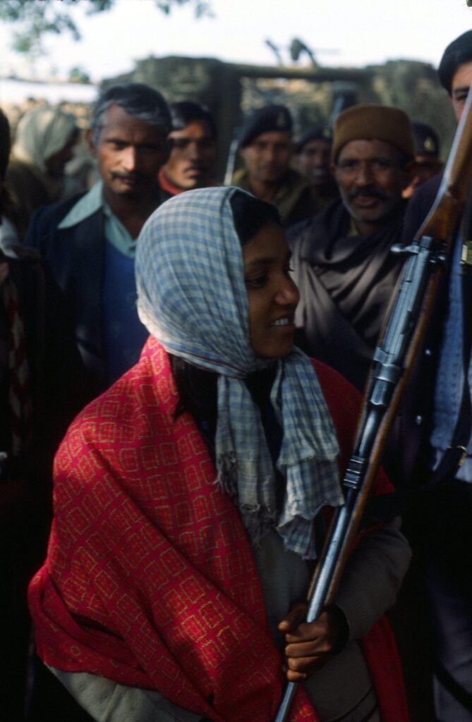Phoolan Devi & her famous gun - 1983 Phoolan Devi in a red shawl and checked head scarf holding her rifle, surrounded by a crowd of men, 1983